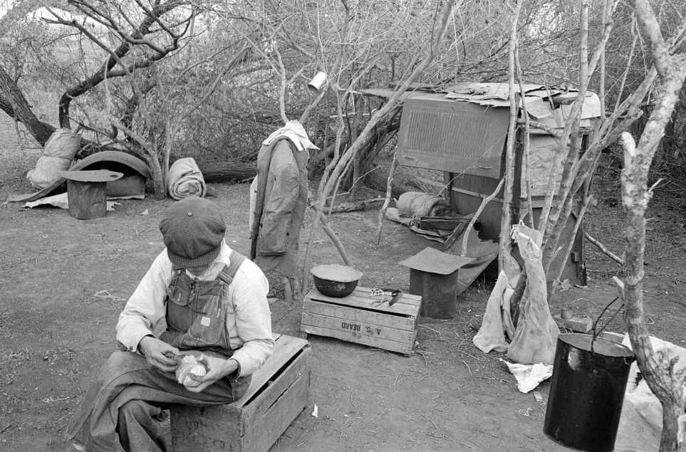 Migrant worker living in camp with two other migrant men. His sleeping quarters and all worldly possession are to be seen in background. Harlingen, Texas Photo: Russell Lee