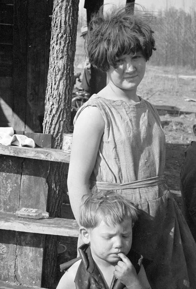 Twelve-year old girl of family of nine living in one-room hut built over the chassis of abandoned Ford truck in open field on U.S. Route 70 between Camden and Bruceton, Tennessee. Near backward Tennessee section. View also shows one of the small boys in family; the girl is dressed in a meal sack  Photo: Carl Mydans